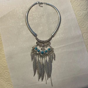Bohemiskt halsband med turkosa stenar och fjädrar - Unikt bohemiskt halsband i silverfärg med detaljerade mönster och turkosa stenar. Halsbandet har hängande fjäderformade detaljer som ger en elegant touch. Perfekt för att lyfta din stil med en bohemisk känsla.