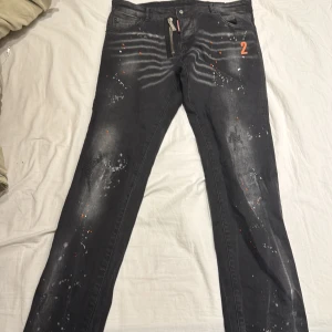 Svarta jeans med färgstänk från Dsquared2 - Snygga svarta jeans från Dsquared2 med unika vita och orange färgstänk. De har en dragkedja framtill och en cool design med nummerdetalj. Perfekta för en trendig look.
