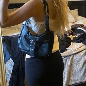 Svart axelväska med spänne - Snygg svart axelväska i skinn med ett stort dekorativt spänne framtill. Perfekt för att bära med sig det viktigaste med stil. Väskan har en praktisk storlek och en justerbar axelrem.