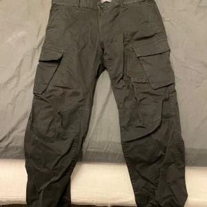 Svarta cargobyxor från Dressmann - Snygga svarta cargobyxor från Dressmann med flera praktiska fickor. Byxorna har en knapp och dragkedja framtill och är perfekta för en avslappnad stil. Regular fit ger en bekväm passform.