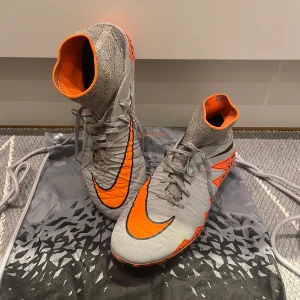 Nike Hypervenom Elite SG - Snygga fotbollsskor från Nike i grått och orange med hög ankel och snörning. Skorna har en iögonfallande design med svarta detaljer och en mönstrad sula för bra grepp på planen. Inga hål eller separationer och har använts sparsamt. OBS! Inga innersulor finns.