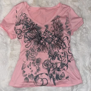 Rosa t-shirt med tryck från SHEIN - Säljer en rosa t-shirt från SHEIN med ett coolt svart tryck av vingar och mönster på framsidan. T-shirten har en v-ringad hals och korta ärmar. Perfekt för en avslappnad stil. Aldrig använd.