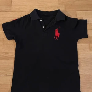 Svart pikétröja med polo loga - Snygg svart pikétröja från Ralph Lauren med en stor röd broderad logga på bröstet. Tröjan har korta ärmar och en klassisk krage med knappar. Perfekt för en stilren look.