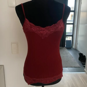 Röd ribbad linnetopp med spetsdetaljer - Snygg röd linnetopp med ribbad struktur och spetsdetaljer vid halsringning och nederkant. Perfekt för en stilren look.