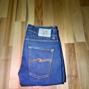Mörkblåa jeans från Nudie Jeans, 31/32 - Säljer ett par snygga mörkblåa jeans från Nudie Jeans. De har en klassisk femficksdesign med orange sömmar och en diskret logga på bakfickan. Perfekta för en stilren look. Riktigt fet tvätt, storlek 31/32, kan finnas små fläckar som knappt syns, jeansen har lagats som ni ser på sista bilden, samt en liten del av bakfickan. Model: Lean Dean