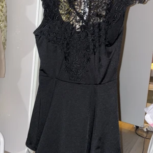 Svart blus med spetsdetaljer - Elegant svart blus med vackra spetsdetaljer på överdelen och en söt rosett i ryggen. Klänningen har en smickrande passform och är perfekt för festliga tillfällen.