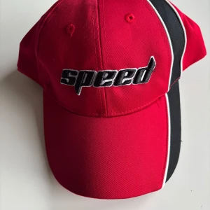 Röd och svart keps med texten 'speed' - Snygg röd keps med svart och vit detalj på sidan och texten 'speed' broderad framtill. Perfekt för en sportig look.