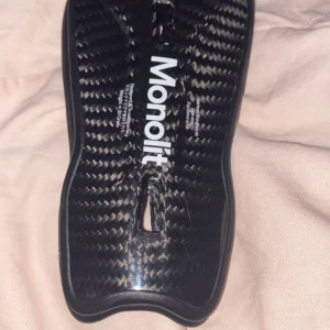 Svarta benskydd från Monolit - Säljer ett par svarta benskydd från Monolit med en stilren design. De har en kolfiberliknande yta och är formade för att ge bra skydd och komfort. Perfekta för fotboll eller andra sporter där skydd behövs.