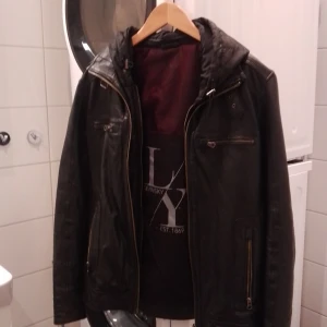Svart skinnjacka med huva - Snygg svart skinnjacka från Levinsky med avtagbar huva och dragkedjor. Jackan har flera praktiska fickor med dragkedja och en stilren design. Perfekt för en cool och avslappnad stil.