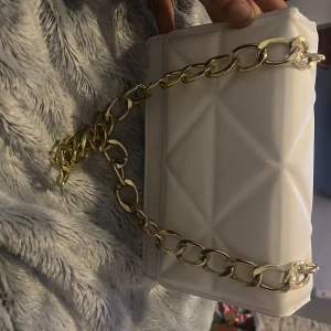 Vit axelväska med kedja - Snygg vit axelväska med quiltad design och en elegant guldkedja som axelrem. Väskan har en stilren look med magnetknappar för enkel öppning och stängning. Perfekt för att ge din outfit en chic touch.