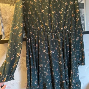 Grön blommig klänning - Charmig grön klänning med blommigt mönster. Den har långa ärmar och en bekväm passform som passar perfekt för både vår och höst. Klänningen har en lätt rynkad midja som ger en fin siluett.