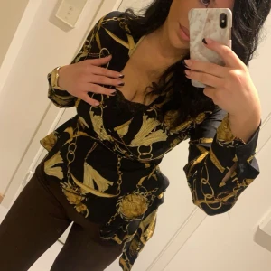 Svart och guldmönstrad blus - Snygg svart blus med guldmönster och omlott-design. Blusen har långa ärmar och ett knytband i midjan för en smickrande passform. Perfekt för en elegant look. Använd en gång