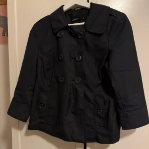 Svart trenchcoat med knappar - Snygg svart kappa med dubbelknäppning och trekvartsärmar. Jackan har en klassisk krage och dekorativa knappar på baksidan. Perfekt för höst och vår.