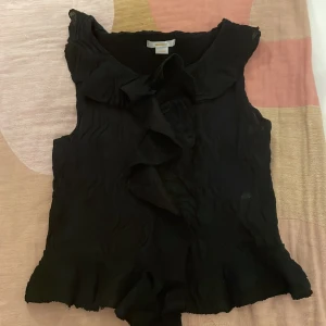 Svart ärmlös blus från Gina Tricot - Snygg svart ärmlös blus från Gina Tricot med volangdetaljer framtill. Perfekt för en stilren look. Tillverkad i ett lätt och luftigt material.