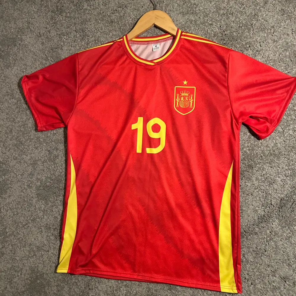 Snygg röd fotbollströja med gula detaljer och nummer 19 på både fram- och baksidan. Tröjan har korta ärmar och ett emblem på bröstet. Perfekt för fotbollsfans som vill visa sitt stöd.. T-paidat.