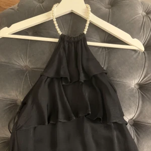 Svart halterneckklänning med pärldetaljer - Elegant svart halterneckktop med pärlor runt halsen. Toppen har volanger framtill och en luftig passform, perfekt för festliga tillfällen.