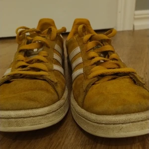 Adidas Campus sneakers i senapsgul mocka - Säljer ett par Adidas Campus sneakers i senapsgul mocka med vita ränder. Skorna har en klassisk design med snörning och är i bra skick, men med lite slitage på sulorna. Perfekta för vardagsbruk och ger en retro känsla till din outfit.