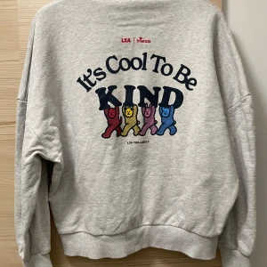 Grå hoodie med tryck från LXA - Supermysig grå hoodie från LXA med ett färgglatt tryck på ryggen som säger 'It's Cool To Be Kind'. Framsidan har en liten gul figur som ger en extra touch. Perfekt för chilliga dagar eller när du vill sprida lite positivitet. Den har en klassisk passform med ribbade muddar och en skön huva. Använd ett fåtal gånger så i väldigt bra skick! Nypris över 1000kr.