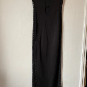 Svart långklänning med spetsdetaljer - Snygg svart långklänning med unika spetsdetaljer längs sidorna. Klänningen har en tight passform och coola utskärningar framtill som ger en edgy look. Perfekt för festliga tillfällen eller en utekväll. Materialet är mjukt och stretchigt för en bekväm känsla.