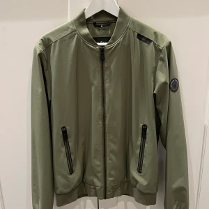 Olivgrön bomberjacka från Sail Racing - Säljer en snygg olivgrön bomberjacka från Sail Racing i storlek medium. Jackan har en dragkedja framtill och två praktiska fickor med dragkedjor. Perfekt för höst och vår, med en stilren design och ribbade muddar vid ärmslut och nederkant.