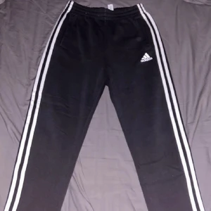 Svarta mjukisbyxor från Adidas - Säljer ett par svarta mjukisbyxor från Adidas i bra skick. De har de klassiska tre vita ränderna längs sidorna och en elastisk midja för extra komfort. Perfekta för träning eller en avslappnad dag hemma. Adidas-loggan är broderad på framsidan. Passar perfekt för höst och vinter! Bra skick