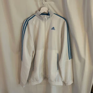 Vit träningsjacka från Adidas - Säljer en snygg vit träningsjacka från Adidas med klassiska blå ränder längs ärmarna. Jackan har dragkedja och två fickor framtill. Perfekt för träning eller som en sportig vardagslook. Den är i mycket bra skick och passar perfekt för vår och höst.