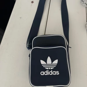Svart axelväska från Adidas - Snygg svart axelväska från Adidas med det klassiska trefoil-loggan i vitt. Väskan har en justerbar axelrem och en praktisk dragkedja upptill. Perfekt för vardagsbruk eller när du är på språng. Den är både stilren och funktionell!