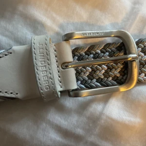Flätat bälte från J.Lindeberg - Säljer ett snyggt flätat bälte från J.Lindeberg i grå och vita toner. Bältet har en stilren silverfärgad spänne och detaljer i vitt skinn. Perfekt för att ge en stilren touch till din outfit. Passar både till vardag och fest! Original pris 928 kr.