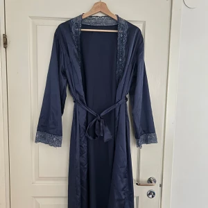 Mörkblå morgonrock med spetsdetaljer - Säljer en elegant mörkblå morgonrock med vackra spetsdetaljer längs kragen och ärmsluten. Den har ett knytband i midjan för en justerbar passform. Perfekt för mysiga morgnar eller kvällar hemma. Materialet känns mjukt och lyxigt mot huden. Storlek L/XL. 