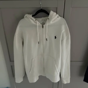 Vit hoodie från Polo Ralph Lauren - Säljer en snygg vit hoodie från Polo Ralph Lauren i storlek M. Den har en dragkedja framtill och en klassisk logga på bröstet. Perfekt för både vardag och chill. Huvan har snören för justering och det finns fickor framtill. Passar perfekt till höst och vår!