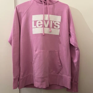 Rosa hoodie från Levi's - Säljer en superfin rosa hoodie från Levi's i mycket bra skick. Den har en stor ficka fram och en justerbar huva med dragsko. Perfekt för en avslappnad stil och passar bra till både jeans och leggings. Stor logga på bröstet ger en cool look!