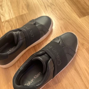 Svarta sneakers från Calvin Klein - Säljer ett par svarta sneakers från Calvin Klein i bra skick. De har en stilren design med elastiska band över foten och en vit sula. Perfekta för både vardag och festliga tillfällen. Skorna är bekväma och lätta att matcha med olika outfits.