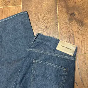 Säljer ett par snygga mörkblå jeans från J.Lindeberg. Storlek 32/32. Onvända. De har en klassisk design med fem fickor och en dragkedja. Perfekta för både vardag och fest! 🕺