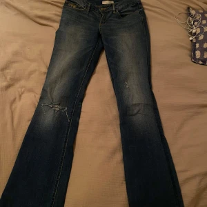 Blå jeans bootcut - Säljer ett par snygga blå jeans i bra skick. De har en tight passform och är logmidjade. Jeansen har en klassisk femficksdesign och är perfekta för både vardag och fest. De har en liten slitning på ena benet och detaljer vid fickorna. 