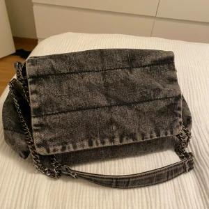 Grå axelväska i jeansmaterial - Säljer en snygg grå axelväska i jeansmaterial med kedjehandtag. Väskan har flera fack och en dragkedja för säker förvaring. Perfekt för både vardag och fest!