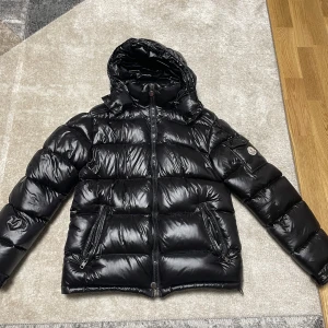 Svart Moncler maya jacka - Säljer en snygg svart pufferjacka från Moncler i mycket bra skick. Jackan är perfekt för vintern med sitt blanka material och varma dunfyllning. Den har en dragkedja framtill och två praktiska fickor med dragkedjor. Huvan är justerbar för extra skydd mot kyla. Perfekt för både vardag och fest!
