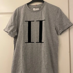 Grå t-shirt från Les Deux - Säljer en stilren grå t-shirt från Les Deux med ett stort svart tryck på framsidan. T-shirten är i normal passform och kortärmad, perfekt för vardagsbruk. Materialet är mjukt och bekvämt och lite ”grövre”, idealiskt för både vår och sommar. Passar bra till jeans eller shorts för en avslappnad look.