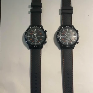 2 st Svarta sportiga klockor med gummiband - Två svarta sportiga klockor med perforerade gummiband och detaljer i orange och vitt. Klockorna har en robust design med tydliga siffror och markeringar på urtavlan. Perfekt för en aktiv livsstil eller som en stilren accessoar.