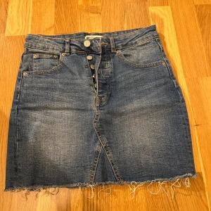 Jeanskjol från ginatricot - Snygg jeanskjol från ginatricot i klassisk blå denim. Kjolen har en rå kant nertill och knappar framtill för en cool look. Perfekt för vardagsbruk eller en avslappnad utekväll. Den har fem fickor och en bekväm passform.