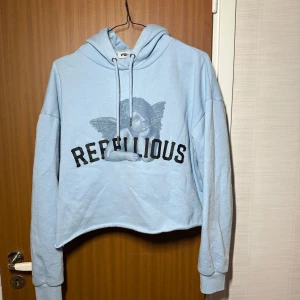 Ljusblå croppad hoodie med tryck - Supercool ljusblå hoodie med texten 'REBELLIOUS' och en ängel på framsidan. Den är croppad och har en skön luva med dragsko. Perfekt för en avslappnad stil. Passar perfekt till jeans, leggings, cargos osv! Hoodien är varm och perfekt för höst och vår!