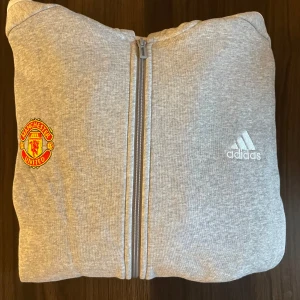 Grå Manchester United hoodie från Adidas - Säljer en grå hoodie från Adidas med Manchester United-logga. Fick Hoodien i julklapp för ett år sedan och hann inte byta in den mot rätt storlek. Aldrig använd! Priset går att diskutera vid en snabb affär.