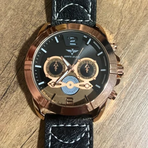 Stilren klocka från Geneva Design - Säljer en snygg klocka från Geneva Design med svart urtavla och roséguldfärgad boett. Klockan har en svart läderrem med vita sömmar som ger en cool kontrast. Perfekt för både vardag och fest! 🕒✨