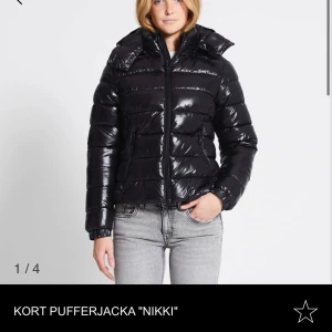 Kort pufferjacka 'Nikki' - Säljer en snygg och trendig kort pufferjacka 'Nikki' i svart. Jackan har en glansig finish och är perfekt för kyliga dagar. Den har en dragkedja framtill och en mysig huva. Passar perfekt till både vardag och fest. Storlek dam xs