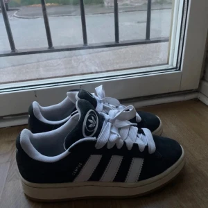 Svarta Adidas Campus sneakers - Säljer ett par snygga svarta Adidas Campus sneakers i mycket bra skick. De har den klassiska tre-randiga designen i vitt och är gjorda i mocka. Perfekta för både vardag och fest! Storleken är tyvärr inte synlig, men de ser ut att vara i standardstorlek.