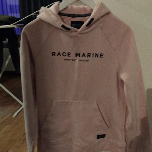 Rosa hoodie från Race Marine - Säljer en supermysig rosa hoodie från Race Marine. Den har en stor ficka fram och texten 'Race Marine' tryckt på bröstet. Perfekt för kyliga dagar eller bara för att chilla hemma. Huvan är skön och ger extra värme. Passar perfekt till jeans eller mjukisbyxor!