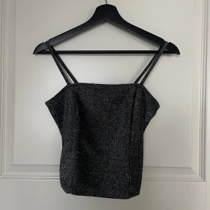 Svart glittrig tubtopp - Snygg svart glittrig tubtopp perfekt för festliga tillfällen. Den har smala axelband och en tight passform som framhäver figuren. Materialet är stretchigt och bekvämt, vilket gör den idealisk för en utekväll. Ett måste i garderoben för alla som älskar att glittra!