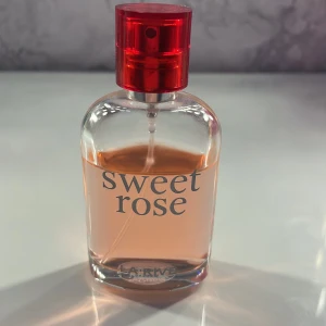 Sweet Rose parfym - Säljer en elegant parfymflaska med doften Sweet Rose från LARIVÉ. Flaskan är genomskinlig med en röd spraykork och innehåller en ljusrosa vätska. Perfekt för dig som älskar blommiga och romantiska dofter. Passar både till vardags och speciella tillfällen. 🌹
