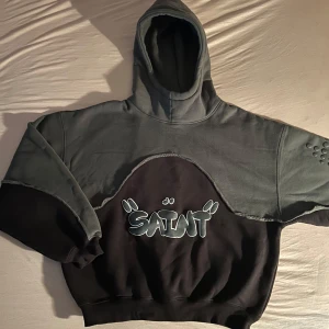 Grå och svart hoodie med texten 'SAINT' - Säljer en cool hoodie i grått och svart med texten 'SAINT' på bröstet. Den har en unik design med detaljer på ärmarna och en bekväm passform. Perfekt för höst och vinter!