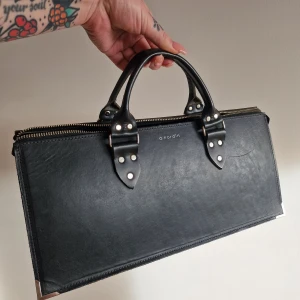 Svart handväska från a.nordin - Snygg och stilren svart handväska i skinn från a.nordin. Väskan har en dragkedja upptill och två robusta handtag med nitar för extra detalj. 