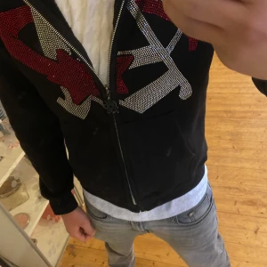 Svart zip från aim for the moon  - Snygg svart hoodie med dragkedja och coola strassdetaljer i rött och silver. Perfekt för att ge din outfit lite extra bling. Den har en bekväm passform och en praktisk huva.